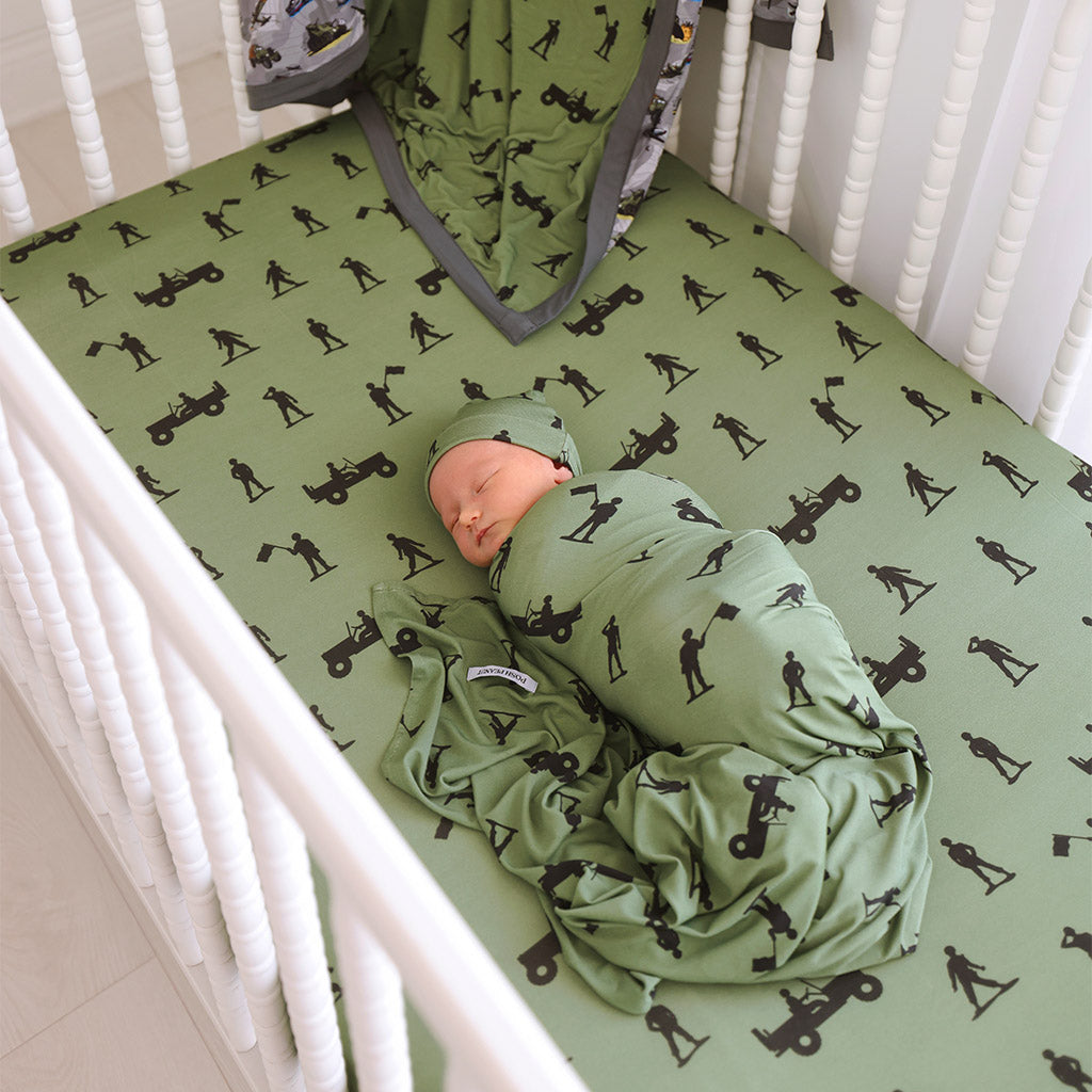 infant wrapped in green army themed swaddle lying in crib with green army print crib sheet