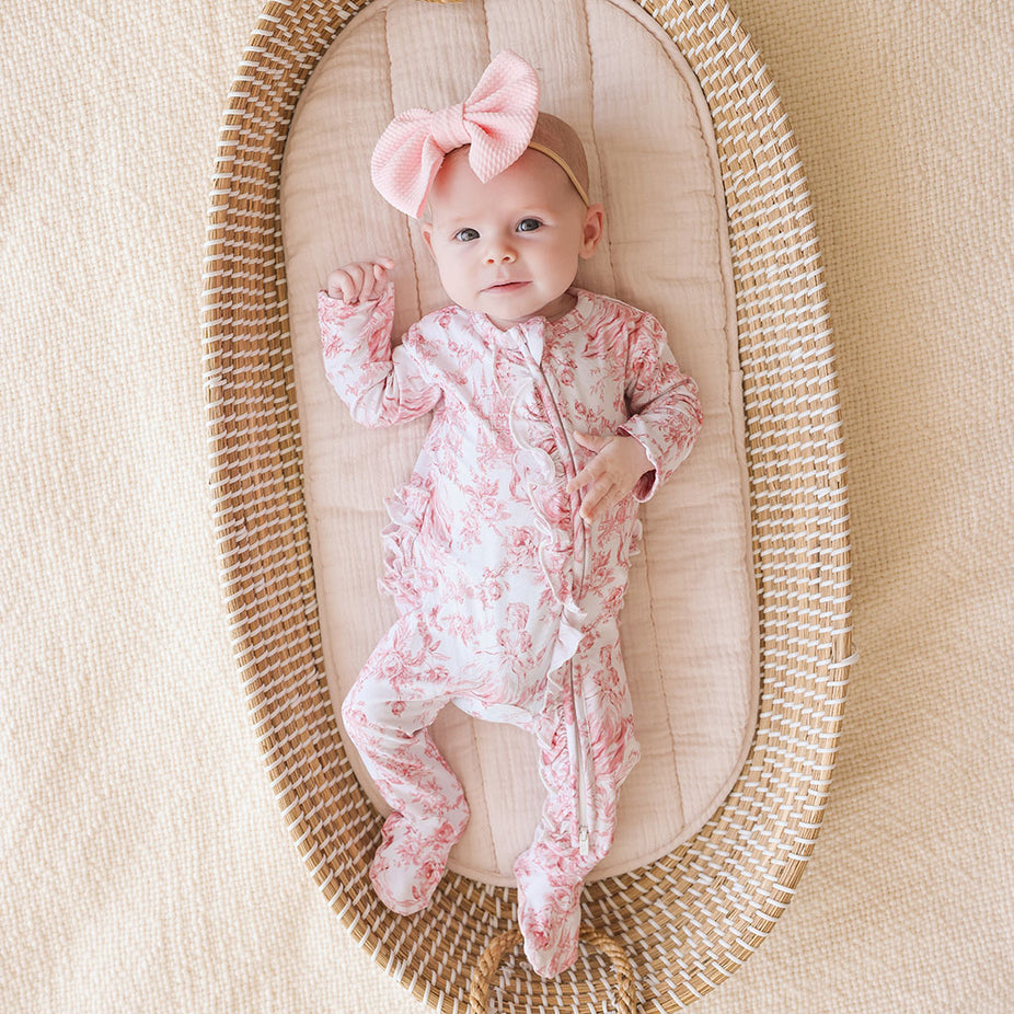 Baby in a floral onesie with a pink bow in a woven basket on a beige background