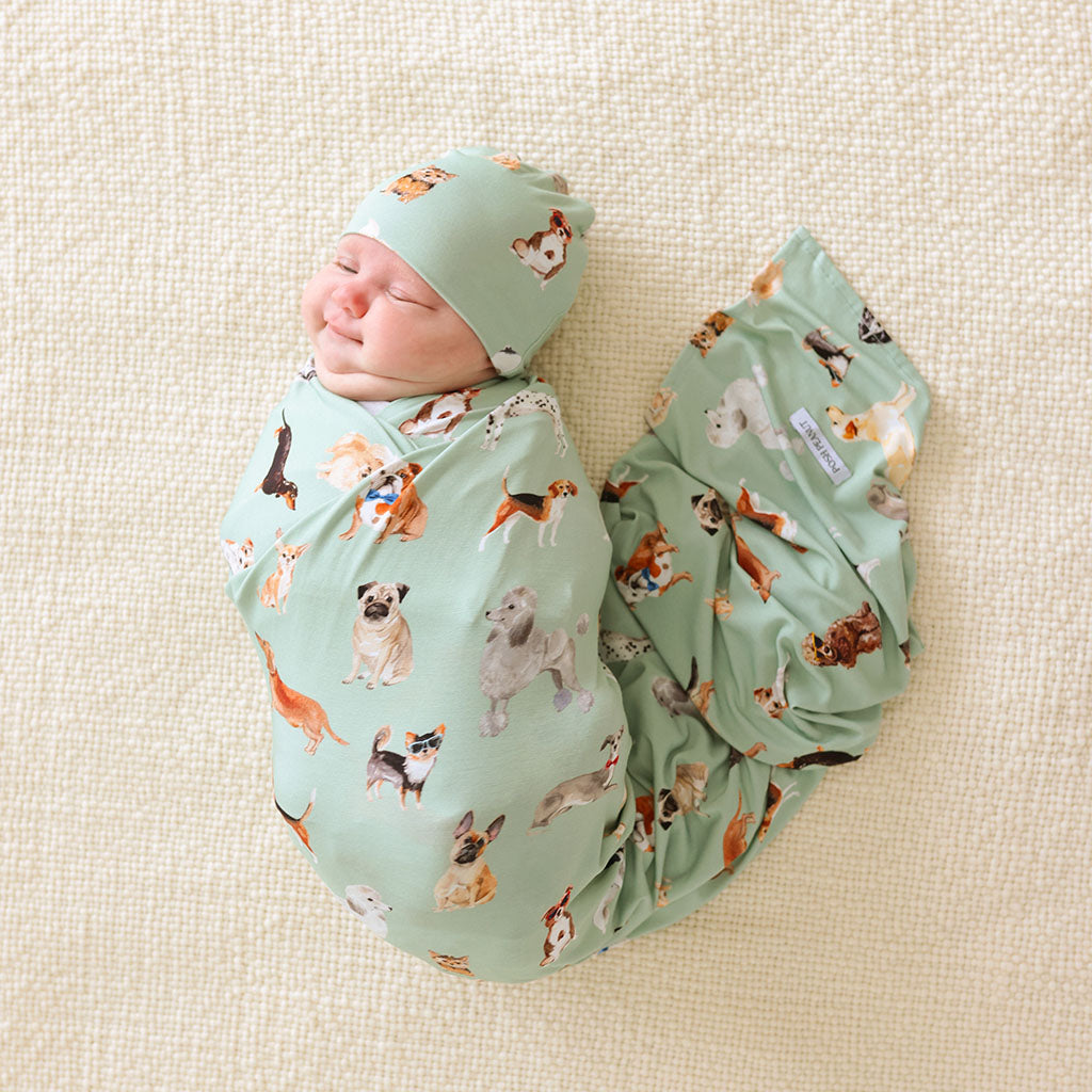 Newborn baby swaddled in a green blanket with matching beanie and dog pattern on a beige surface