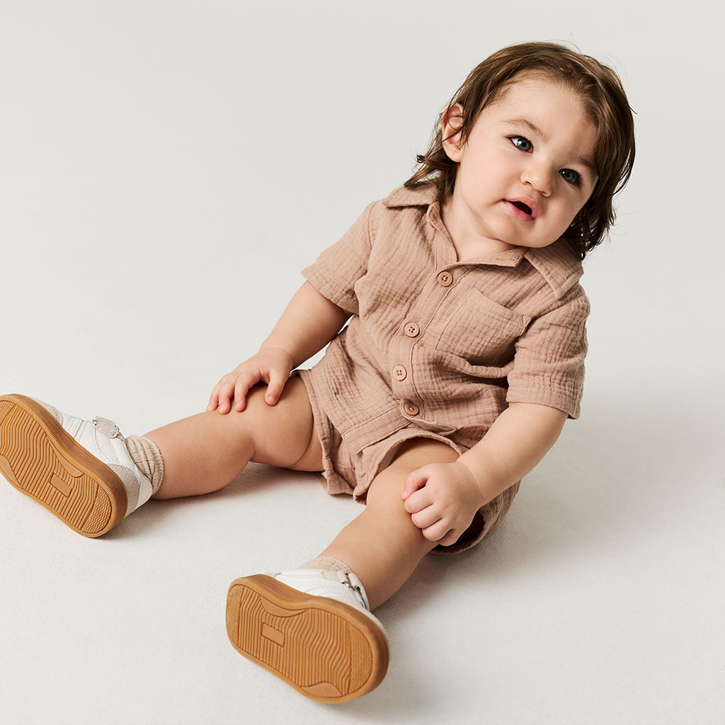 Baby sitting on a white floor wearing a brown outfit with white shoes.