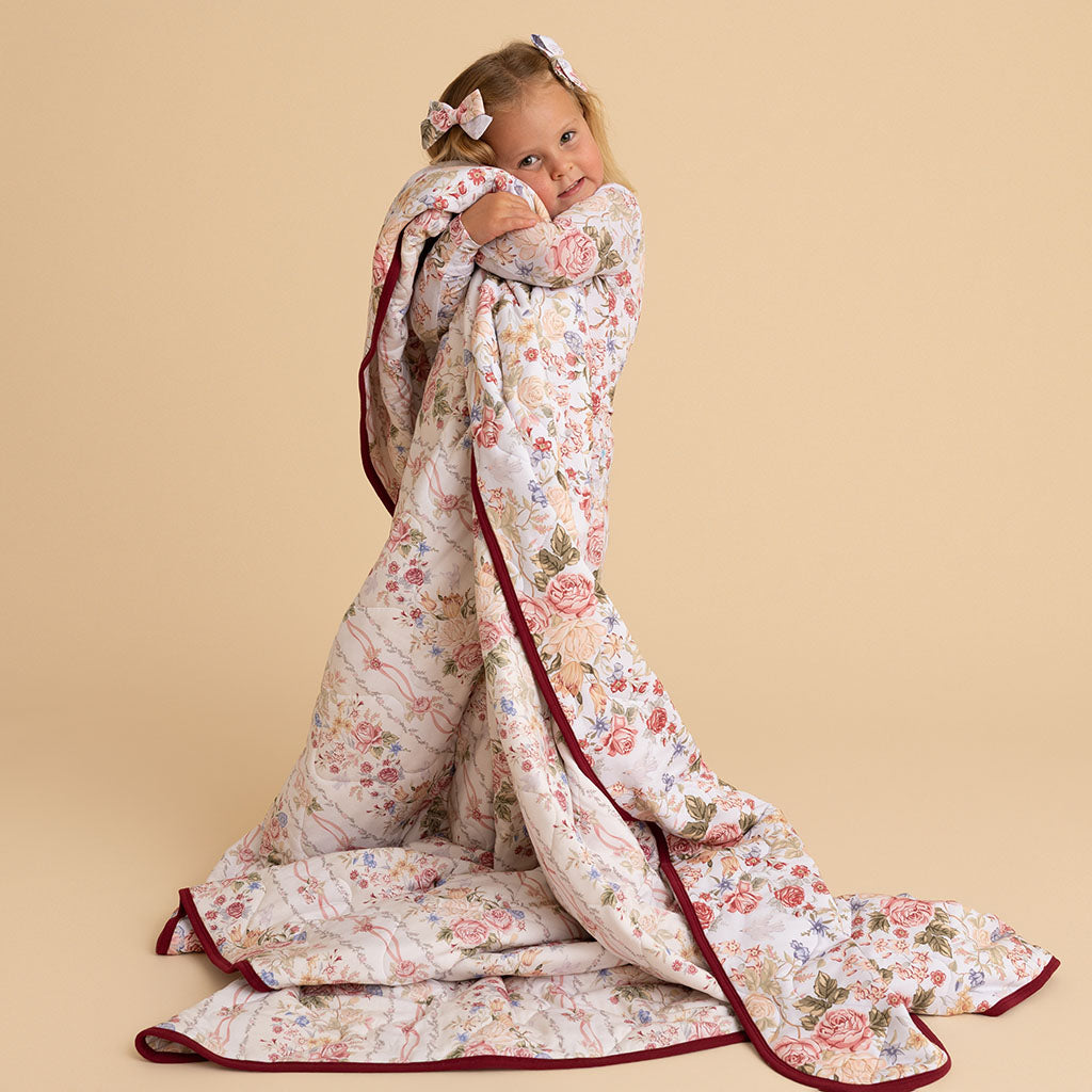 Woman sitting on the floor wrapped in the Reversible Quilted Patoo blanket, the Janey floral print trailing across the floor, on a cream background