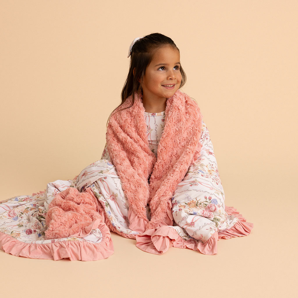 Girl sitting wrapped in the Janey print ruffled Patoo blanket, on a cream background