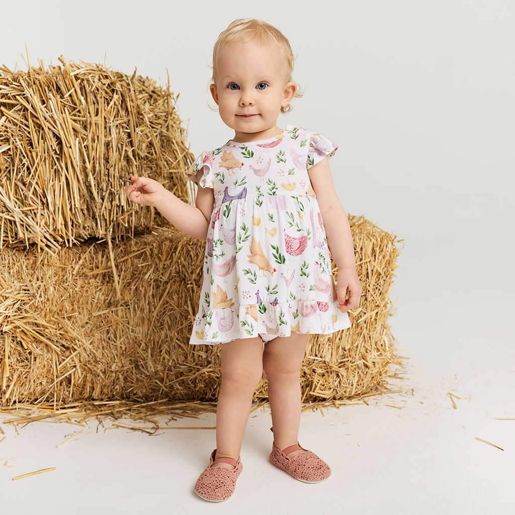 Baby in a floral-chicken print dress standing next to hay bales on a white background