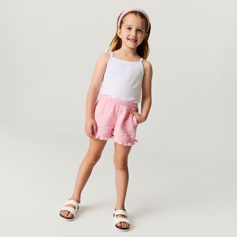 Young girl wearing a white tank top and pink shorts on a plain background