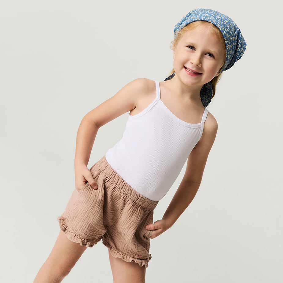 Young girl wearing a white tank top and brown shorts with a blue headscarf on a light gray background