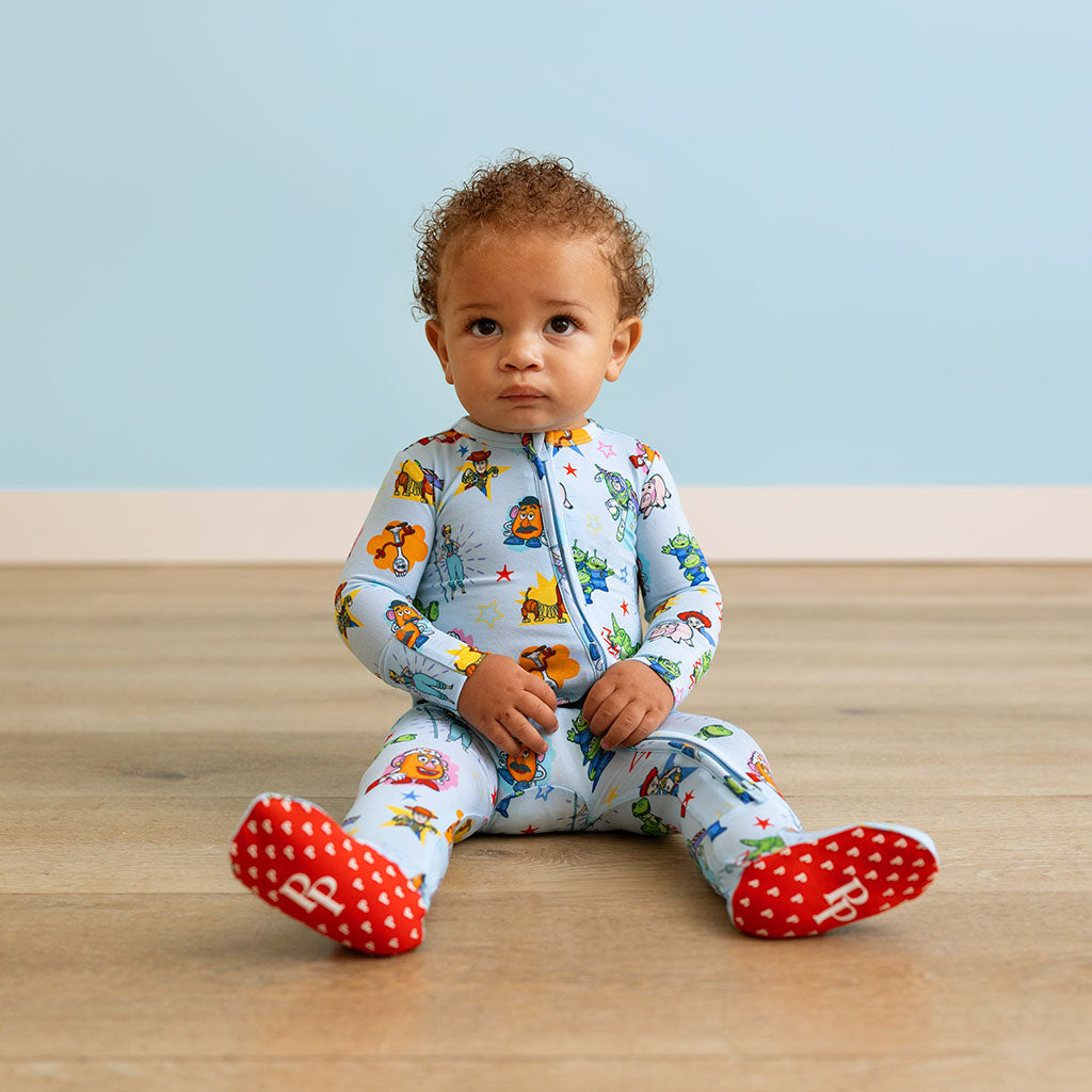 Baby sitting on a wooden floor wearing a colorful onesie with character prints.