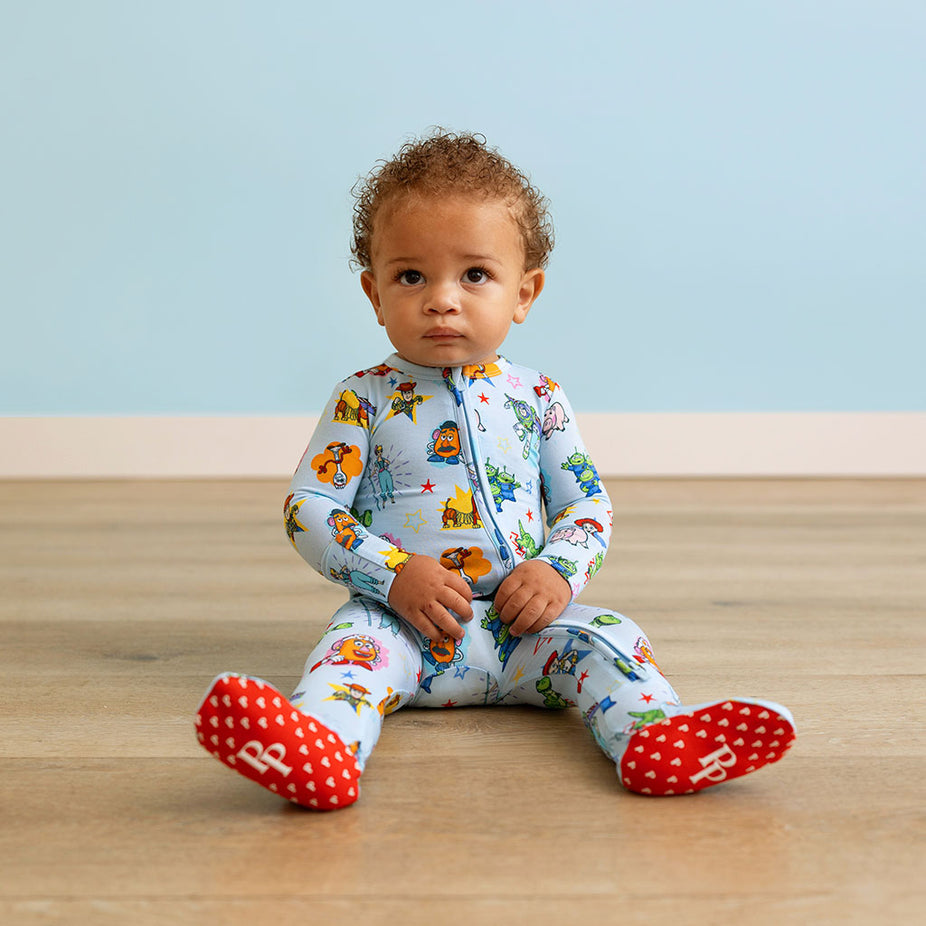 Baby sitting on a wooden floor wearing a colorful onesie with character prints.