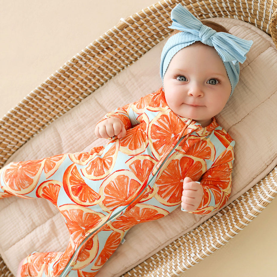 Baby in an orange patterned one piece with a blue headband sitting in a woven basket.