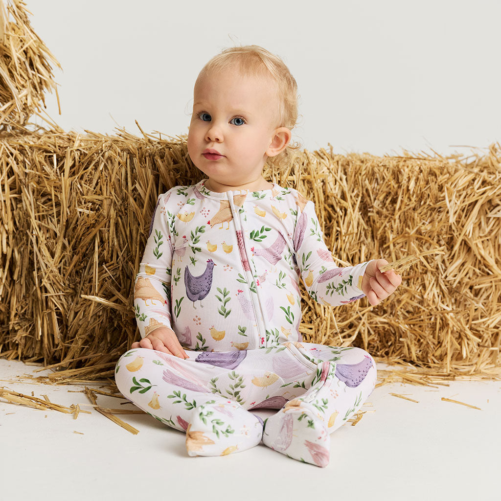 Baby in a floral chicken print one piece sitting on hay bales.