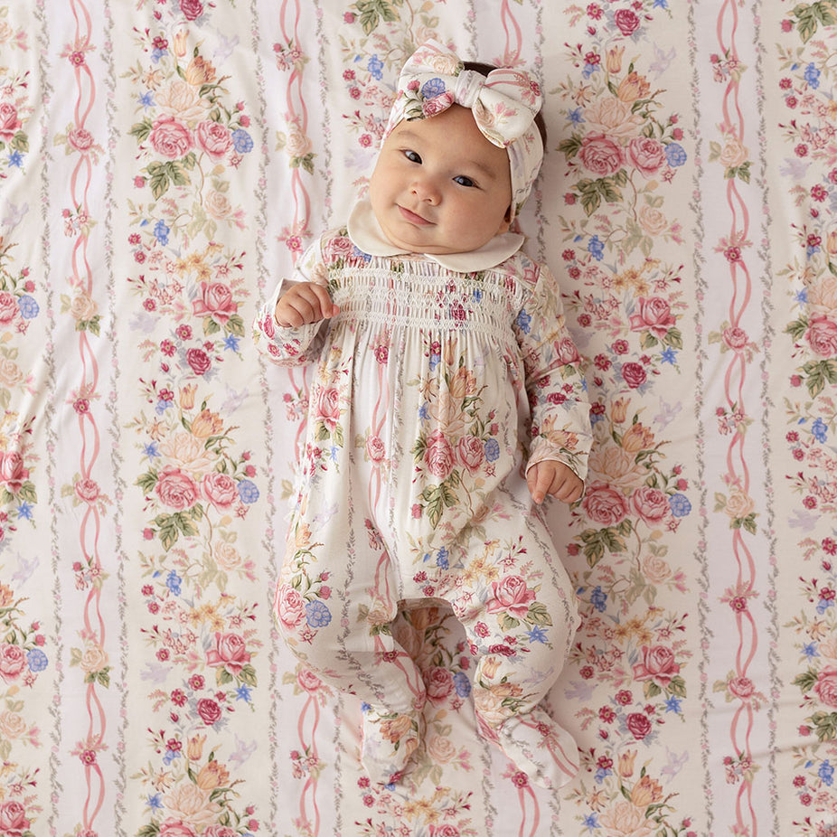 Baby lying on a Janey print fabric backdrop, wearing a matching floral footie with a bow headband