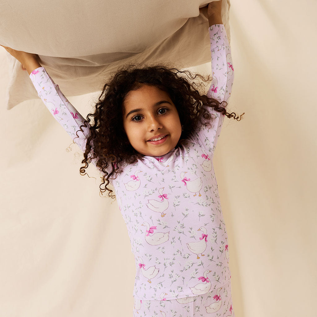 Young girl wearing a light purple bamboo viscose pajama set with a floral goose pattern on a beige background holding a pillow overhead.