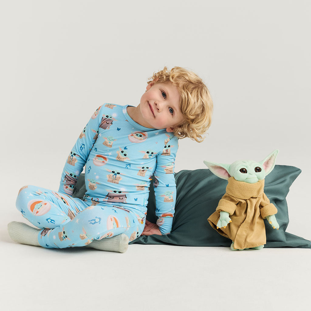 Child wearing a blue pajama set with Star Wars Grogu characters, sitting next to a plush toy resembling 'The Child' from 'The Mandalorian'.
