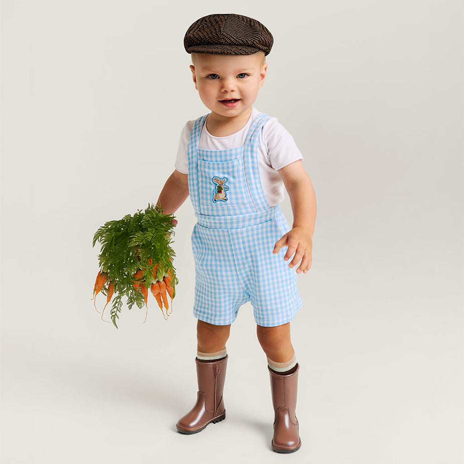 Child wearing a checkered overall with a flat cap, holding carrots on a plain background