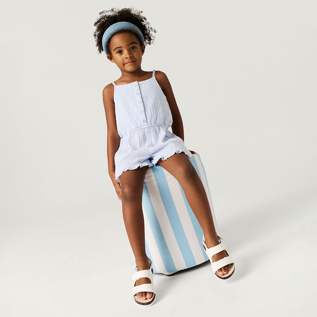 Young girl in a light blue romper sitting on a striped cushion against a plain background