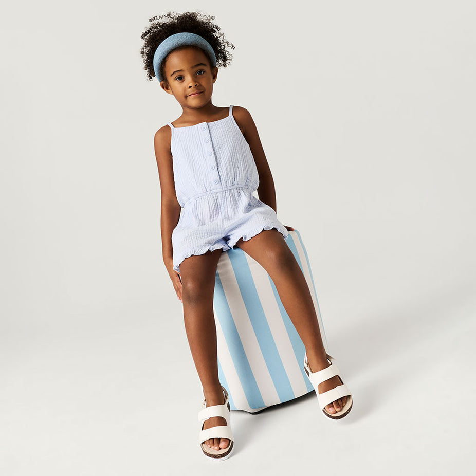 Young girl in a light blue romper sitting on a striped cushion against a plain background