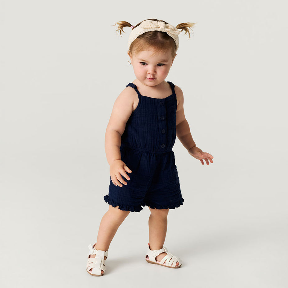 Young child wearing a navy blue romper with white sandals on a light gray background