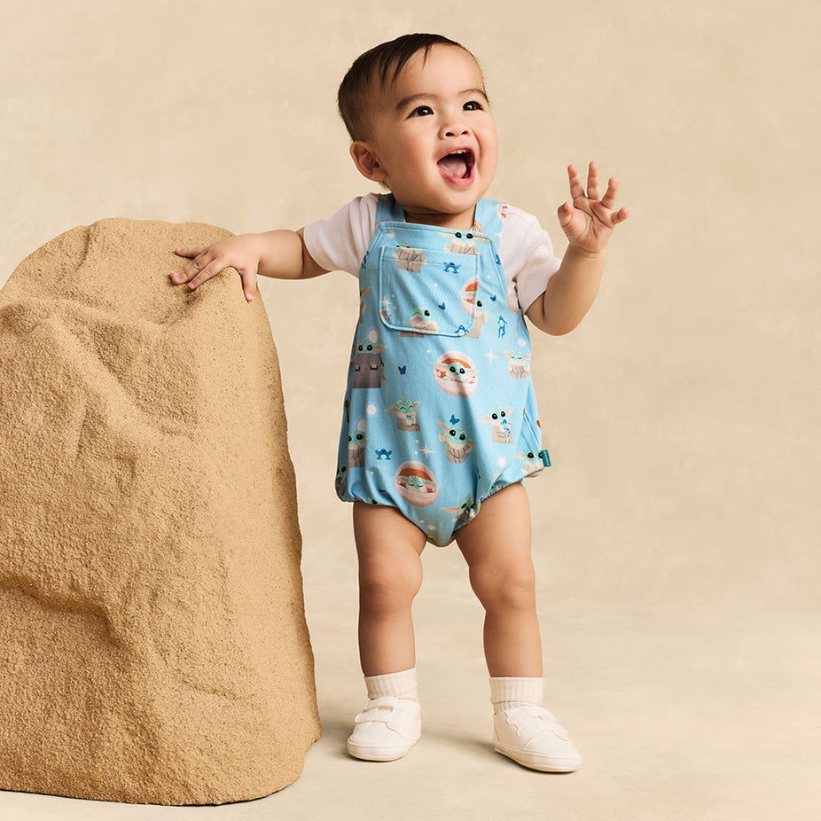 Child standing wearing a blue romper with Star Wars Grogu print against a beige background.