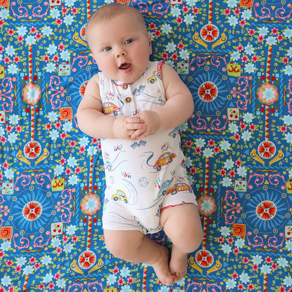 Baby lying on a floral blanket wearing the Posh Peanut Grand Prix Henley Short Romper, a white sleeveless romper printed with colorful cars and road lines.