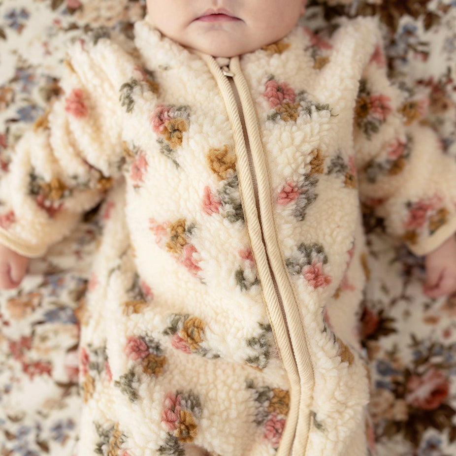 Baby lying down, close-up on face, wearing the Amber print sherpa romper with funnel collar, on a matching floral fabric backdrop