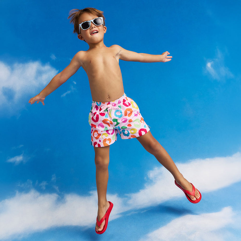 Child in colorful swim trunks and sunglasses jumping against a blue sky with clouds