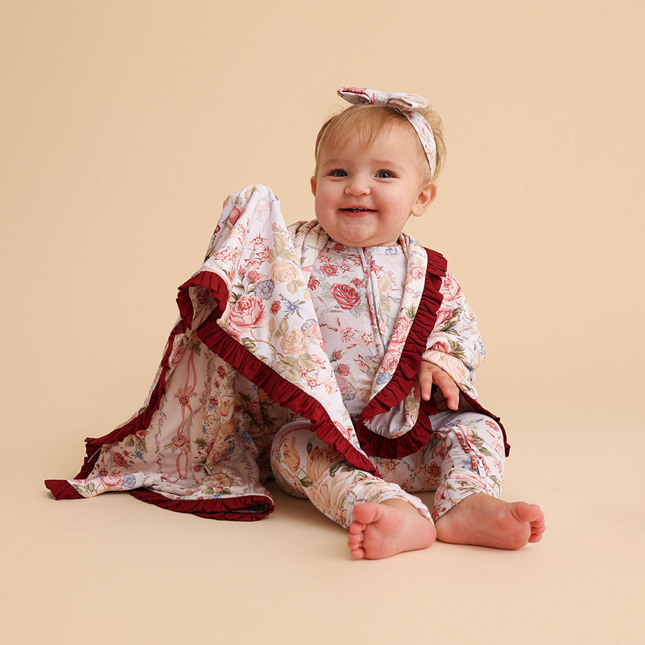 Baby sitting wearing a Janey print outfit, with the Ruffled Luxette Patoo blanket's burgundy ruffle trim visible, on a cream background