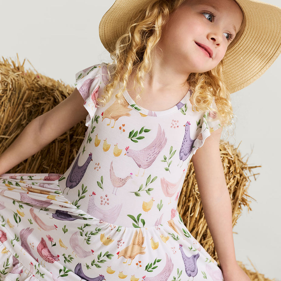 Young girl wearing a floral chicken printed dress and straw hat sitting on hay.