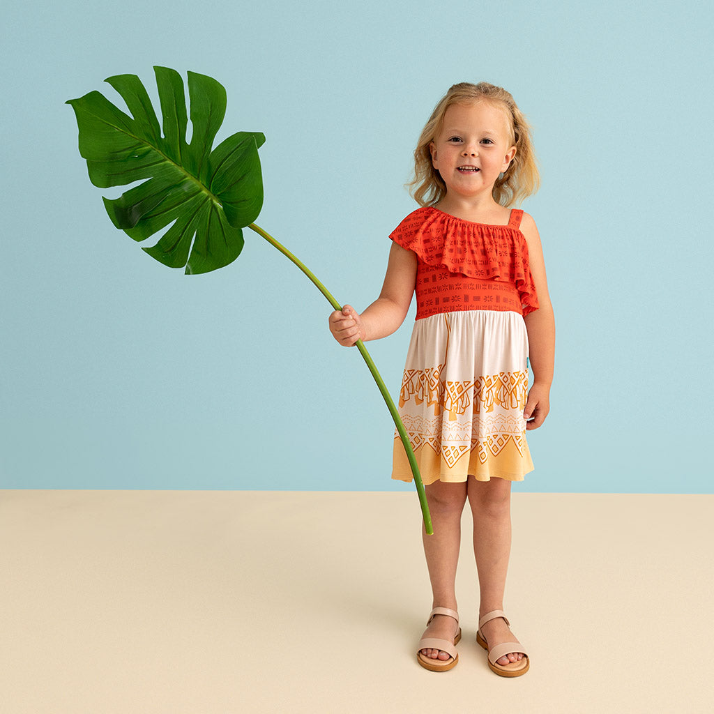 A young girl stands against a light blue and beige studio background, holding a large green monstera leaf. She is wearing a sleeveless tiered dress with a bright coral-red top, cream middle section, and yellow-orange lace trim, paired with tan sandals.