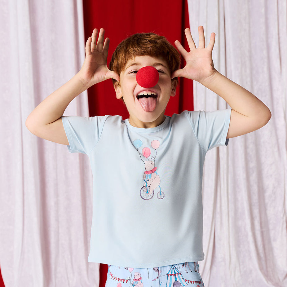 Child wearing a light blue t-shirt with a colorful design and red clown nose, standing against a white curtain.