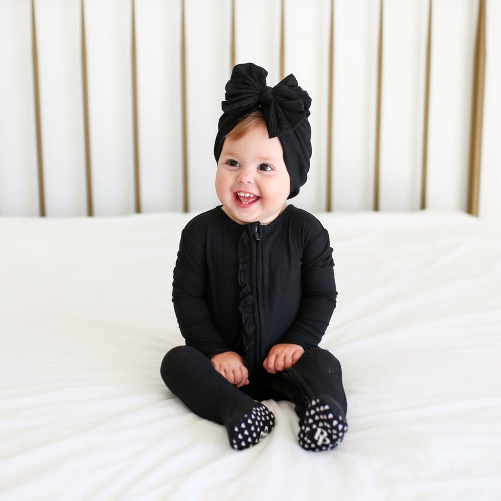 Baby on bed wearing Black Ribbed Footie Ruffled Zippered One Piece