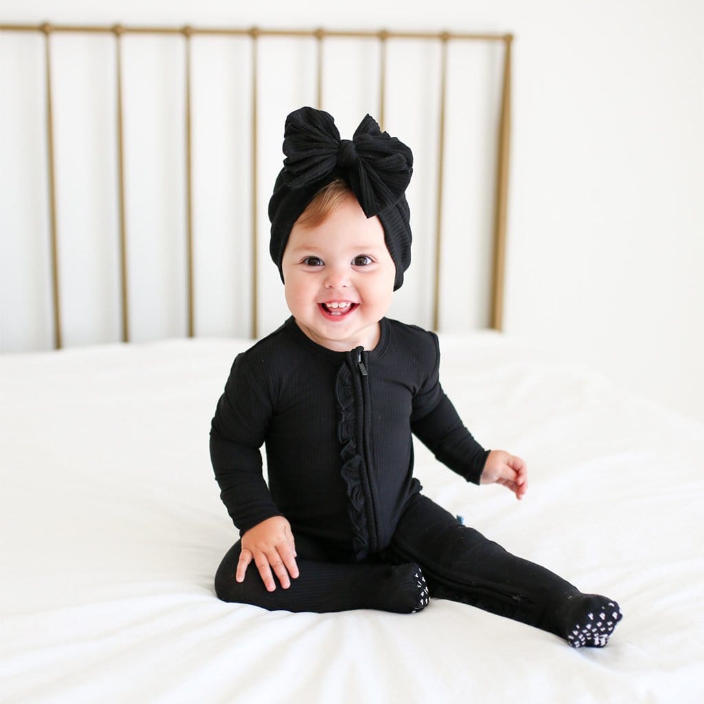 Baby sitting on bed wearing Black Ribbed Footie Ruffled Zippered One Piece